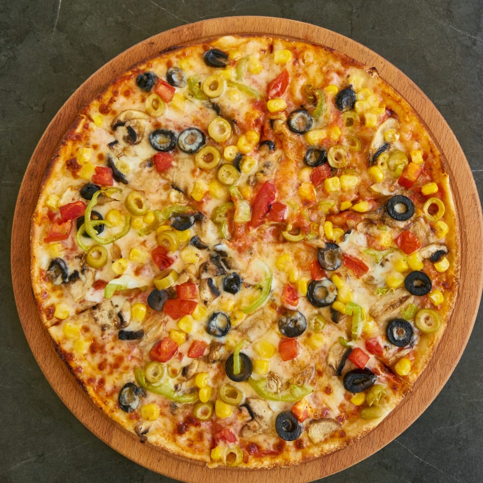 A vibrant vegetarian pizza on a wooden board, topped with olives, corn, peppers, and mushrooms.